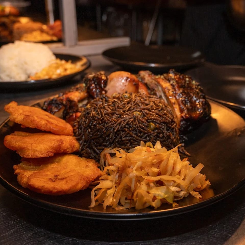 A Haitian meal with dark rice, fried plantains, stewed cabbage, and glazed chicken on a black plate.