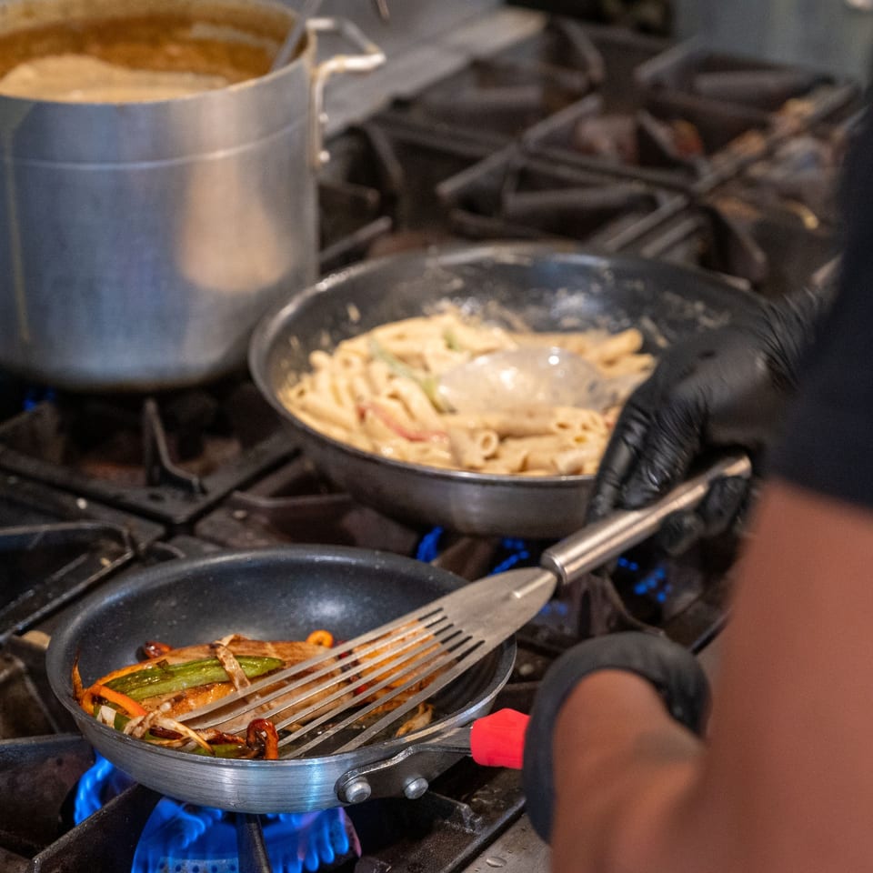 Chef cooking Haitian food, including pasta and vegetables, on a gas stove in a restaurant kitchen.