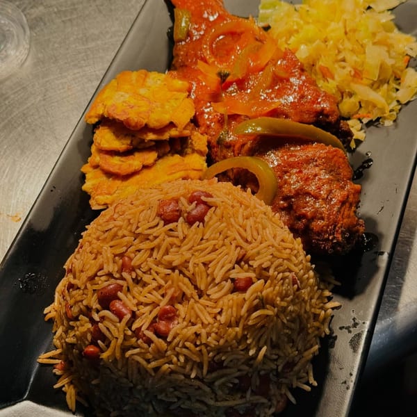 Haitian meal with red snapper, rice and beans, fried plantains, and pikliz on a black plate.