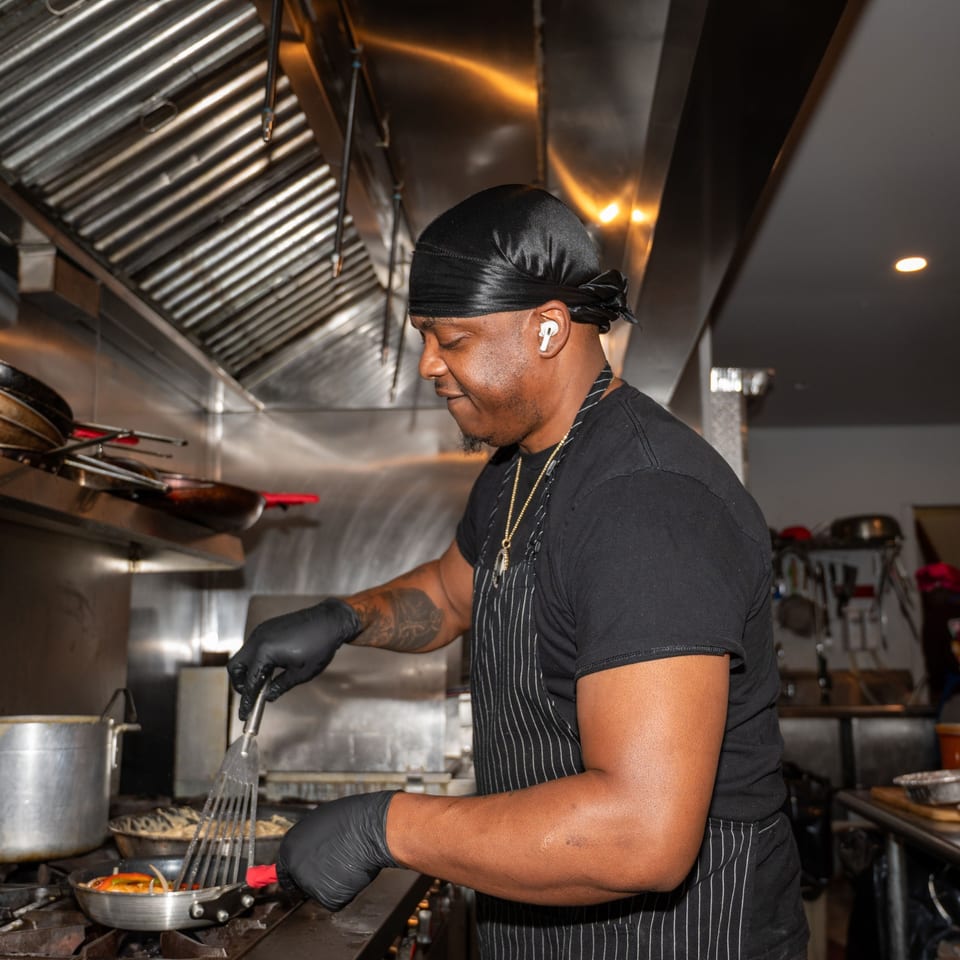 A chef in a black durag and apron cooking Haitian food in a pan on a stove.