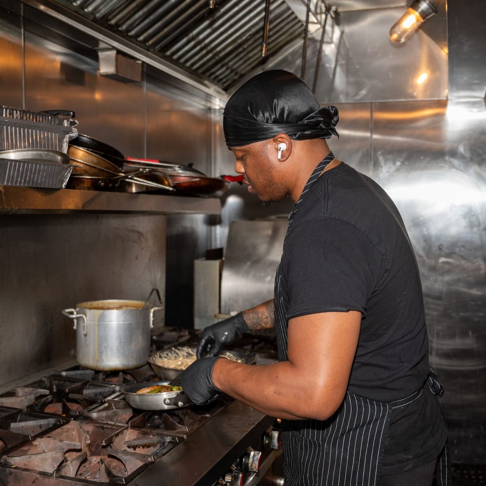 Chef in a black durag and apron cooking Haitian food on a stovetop in a stainless steel kitchen.
